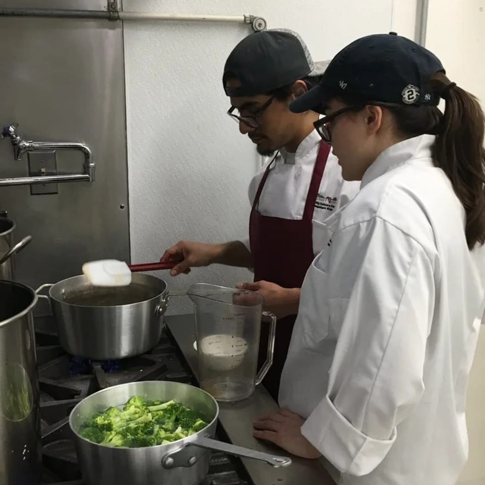 Two chefs prepare food over gas burners, stirring a pot beside a saucepan of broccoli in a commercial kitchen.