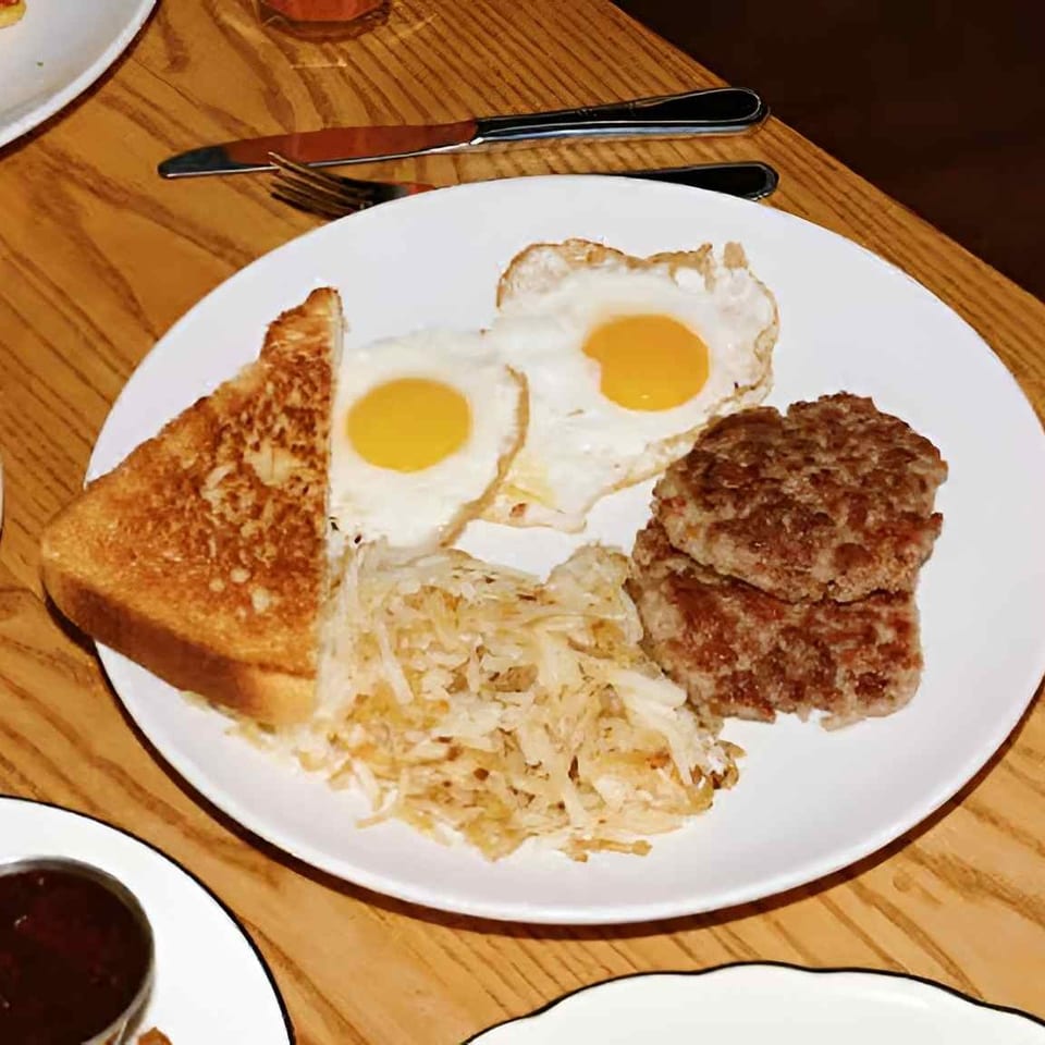 Breakfast plate with two fried eggs, toast, hash browns, sausage patties, and a glass of orange juice on a wooden table.