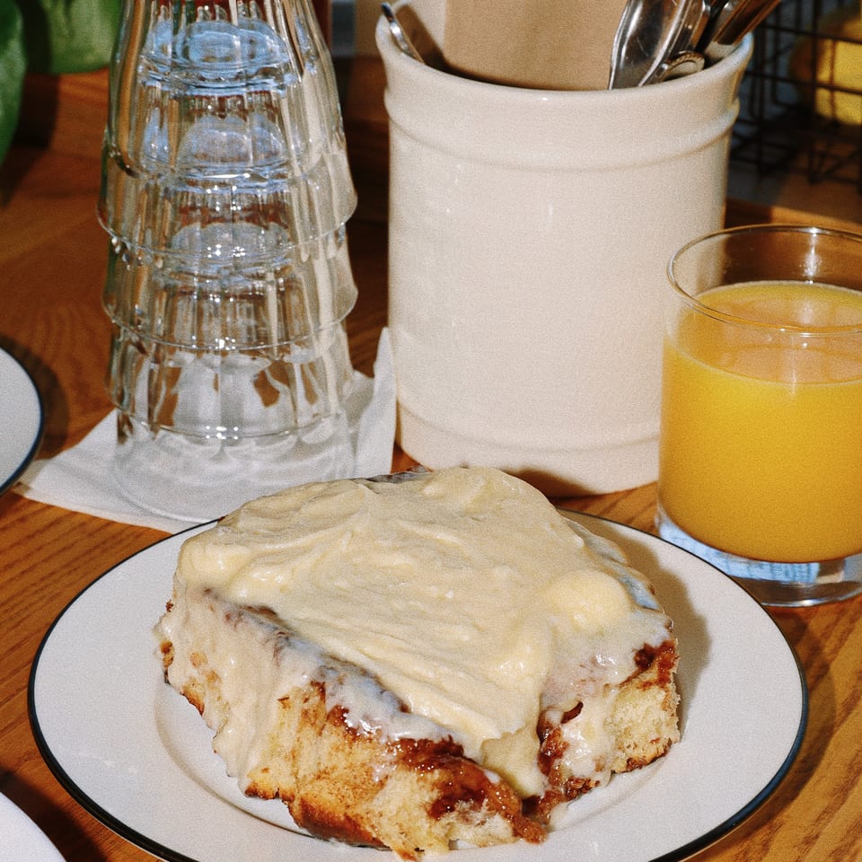 Frosted cinnamon roll on a plate with stacked water glasses, utensils, napkins, and orange juice on a wooden restaurant table.