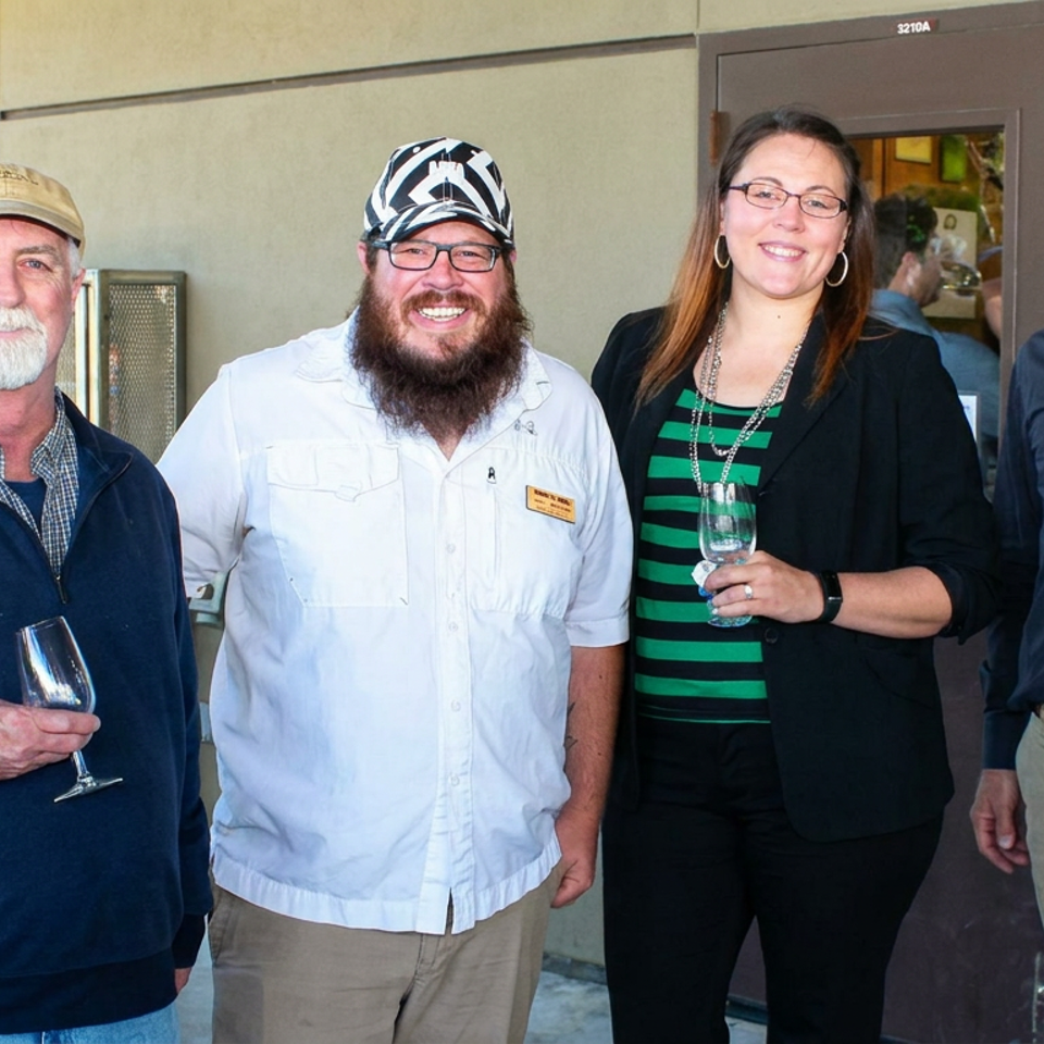 Four adults pose together at a winery entrance, with two holding wine glasses and wooden barrels visible nearby.