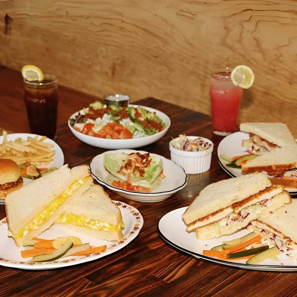 Assorted sandwiches, burger, fries, salads, coleslaw, vegetables, and lime-garnished drinks arranged on a wooden restaurant table.