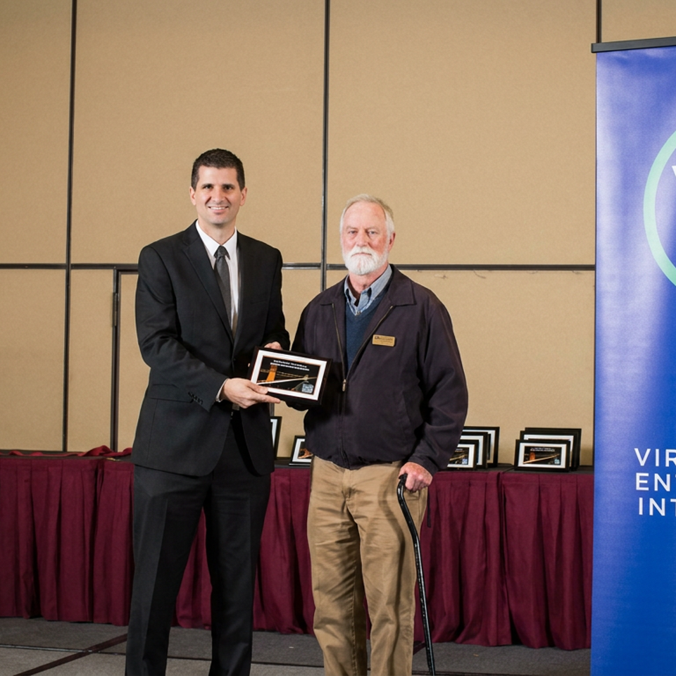 Two men pose with an award beside a Virtual Enterprises International banner and a table displaying framed certificates.