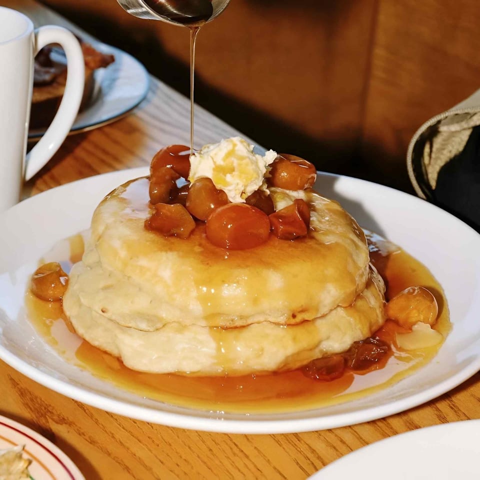 Stack of pancakes topped with whipped cream and strawberries, drizzled with syrup on a plate beside a coffee cup.
